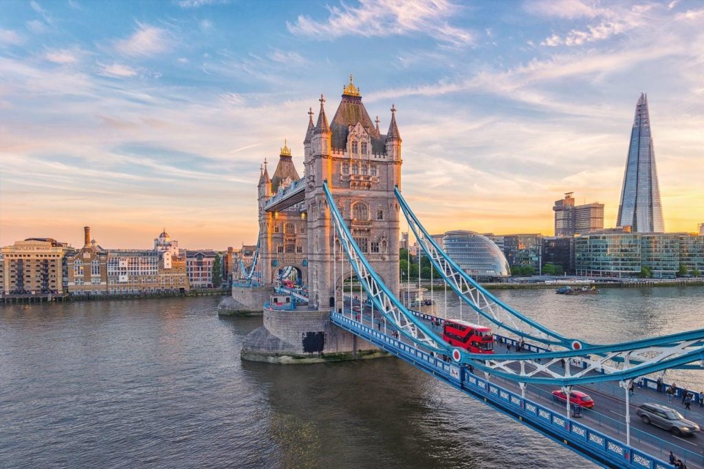 Tower Bridge over the River Thames in London at sunset, with a red double-decker bus crossing and The Shard behind.