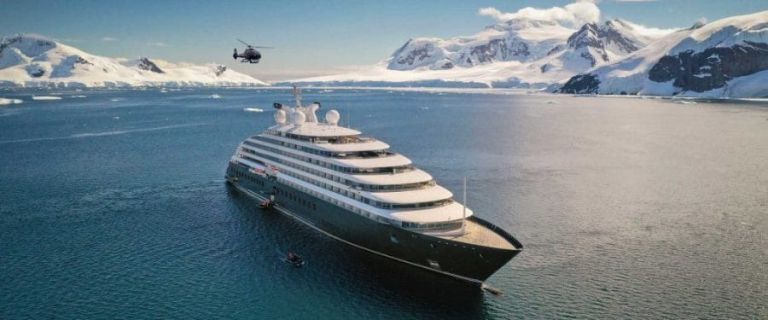 Expedition cruise ship sailing past snow-covered mountains in Antarctica, with a helicopter flying overhead.