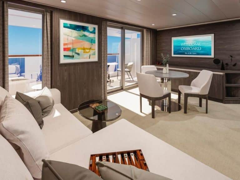 Suite lounge with a white sofa, backgammon board, dining table and wall screen reading 'Welcome Onboard'.