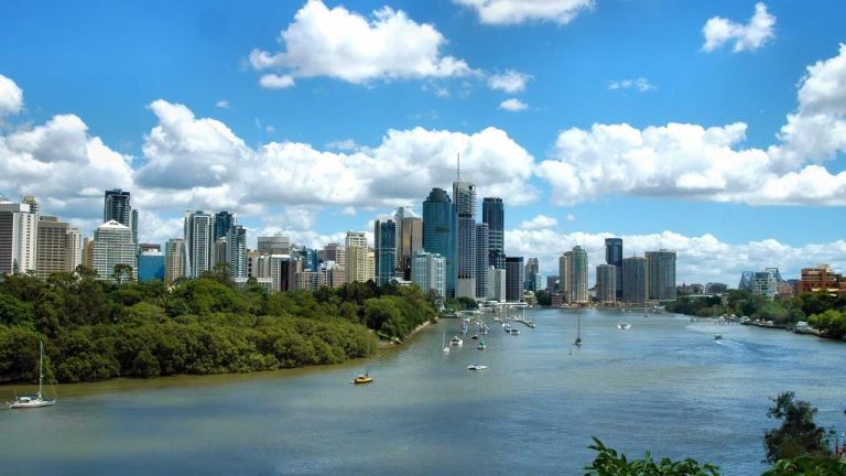 Brisbane skyline across the river, with sailboats on the water and mangroves along the riverbank.