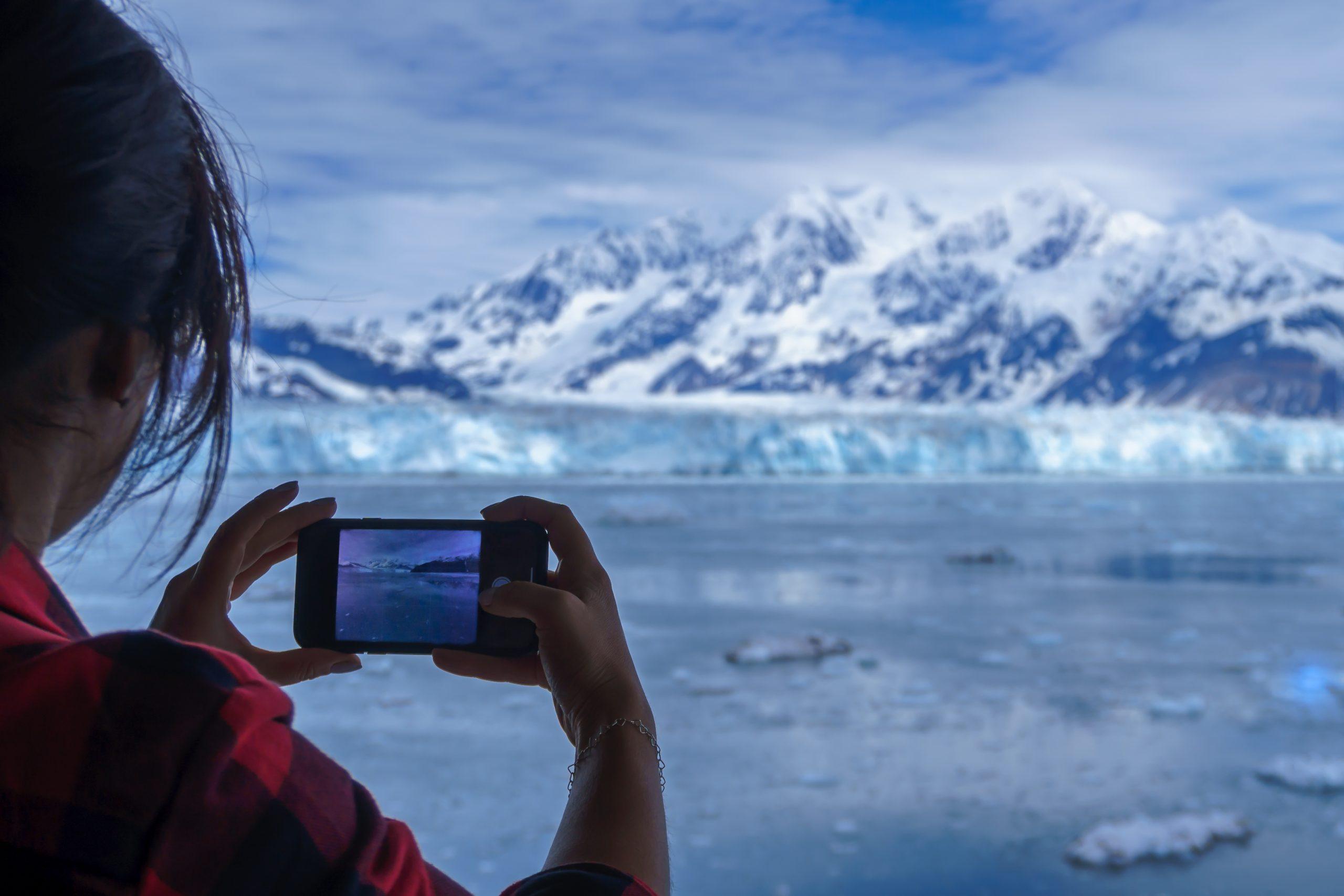 Woman is wearing a lumberjack shirt and taking a picture of Hubbard Glacier on her mobile phone. This is beautiful Alaska and you can see it on the screen of smartphone. Snowy mountains and iceberg.