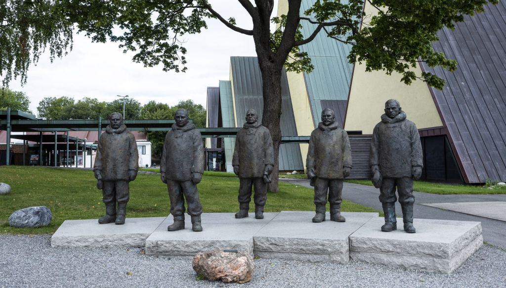 Five bronze statues of polar explorers in heavy parkas on stone plinths in front of a museum building.