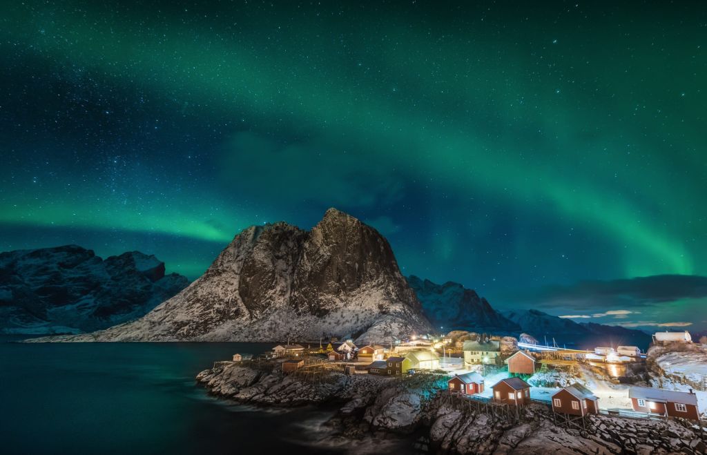 Northern lights over a snowy mountain and a small village of red and yellow fishing cabins on a rocky coast at night.