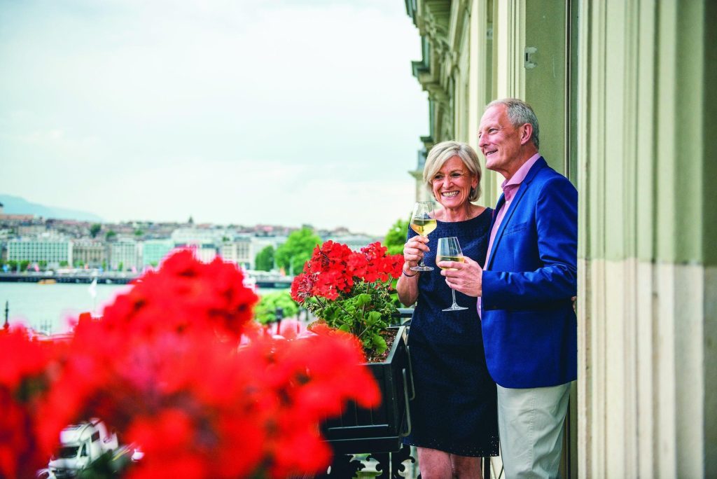 Smiling couple raising wine glasses on a balcony beside red geraniums, with a lake and city below.