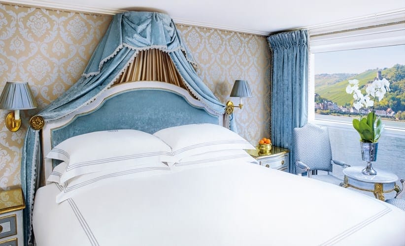 Luxurious hotel bedroom with a blue upholstered headboard, canopy drapes, and white linens by a window with hills outside.