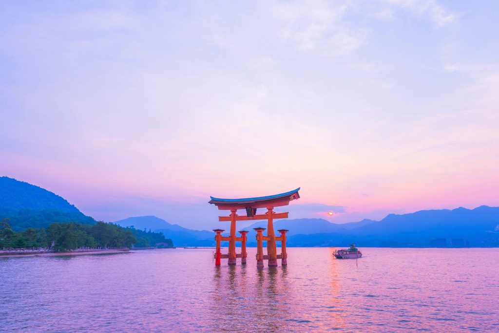 Red torii gate standing in shallow water at sunset, with mountains and a small boat behind.