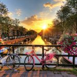 Sunset over an Amsterdam canal with white and pink bicycles on a bridge railing, framed by hanging pink flower baskets.