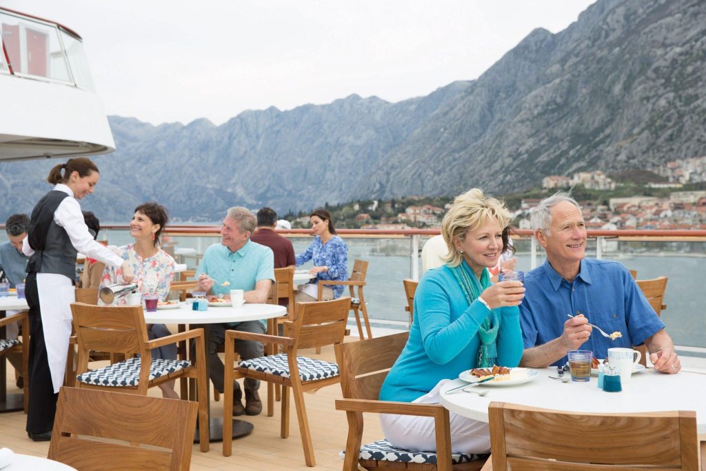 Guests dining on an outdoor deck terrace with a waiter, with mountains and a coastal town behind.