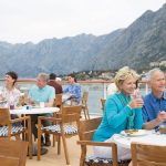 Guests dining on an outdoor deck terrace with a waiter, with mountains and a coastal town behind.