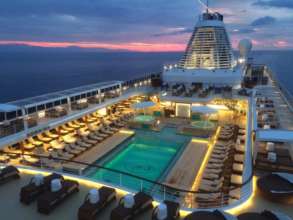 Pool deck at dusk with a glowing blue pool, rows of loungers and two hot tubs under white canopies.