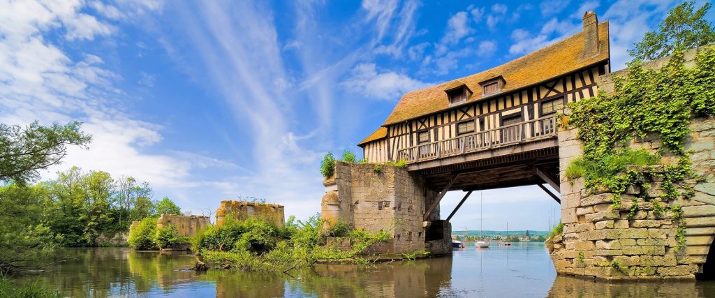 Historic stone bridge with a timber-framed upper level spanning a calm river, ivy-clad walls and a bright blue sky.