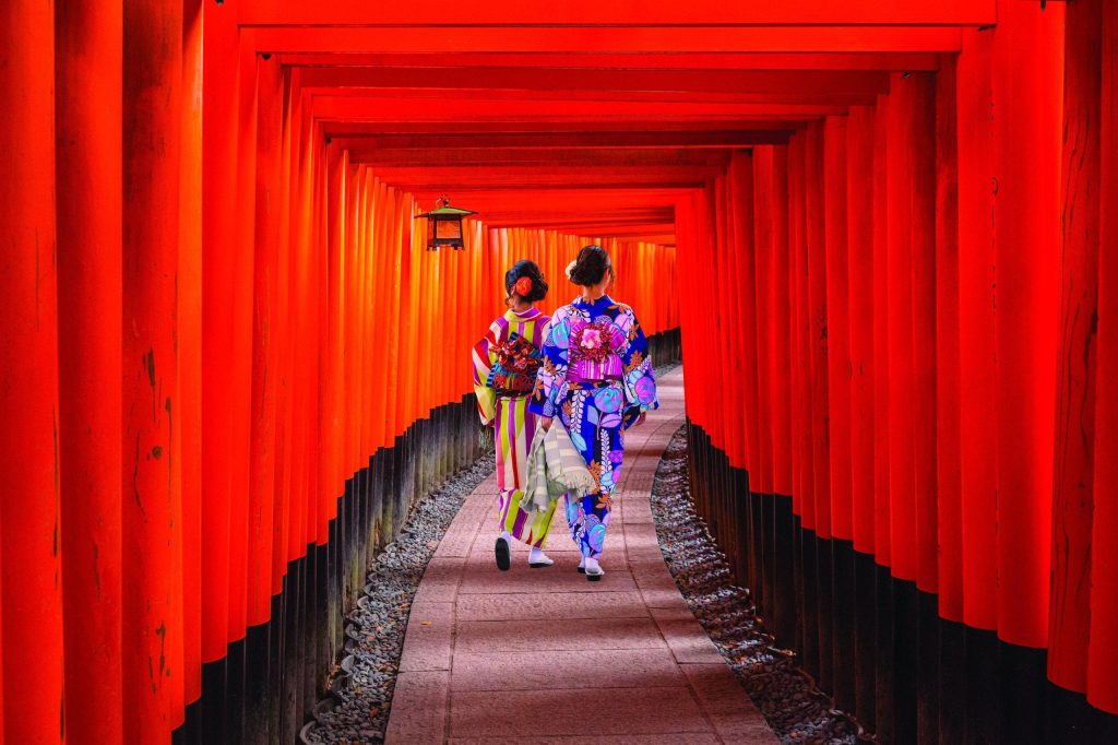 Two women in colourful kimono walking through a tunnel of red torii gates.