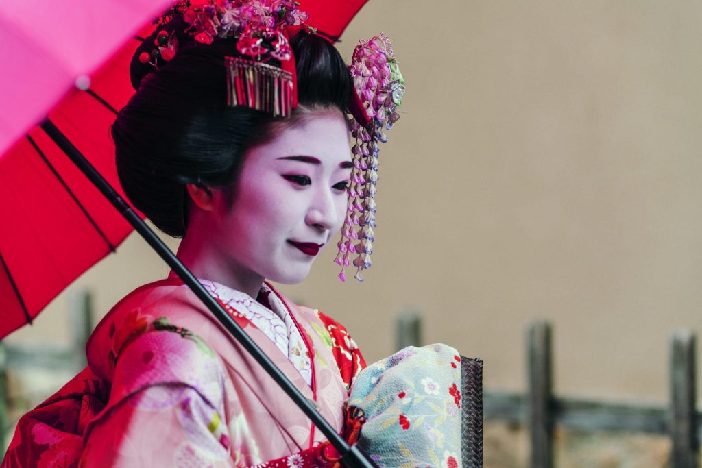 Woman in traditional geisha makeup and kimono holding a red parasol.