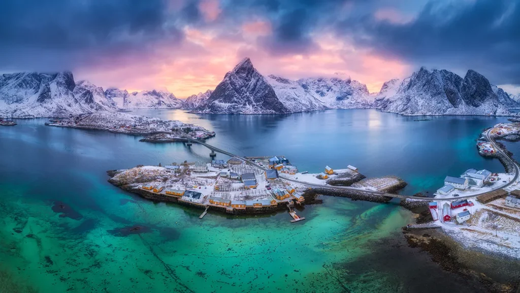 Aerial view of a snowy fishing village on a rocky island with mountains and turquoise water at dusk.
