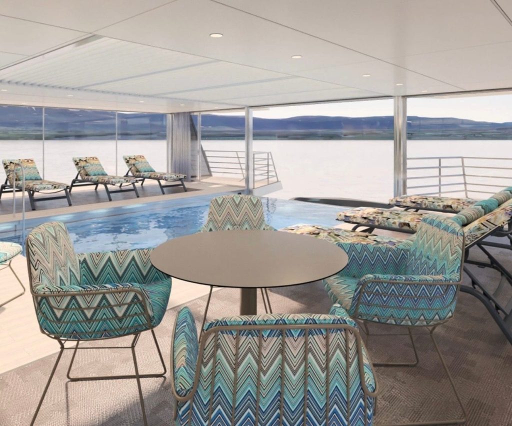 Indoor pool area on a ship or hotel with turquoise chevron chairs around a round table, overlooking a body of water and distant mountains.