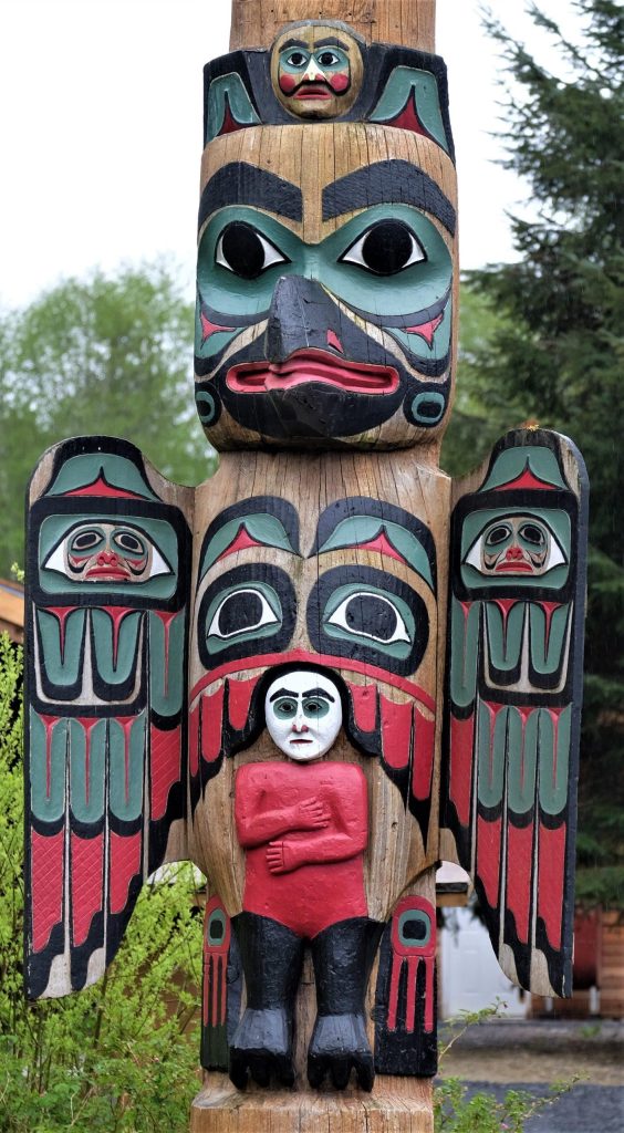 Carved totem pole with red, green and black figures, standing beside trees.