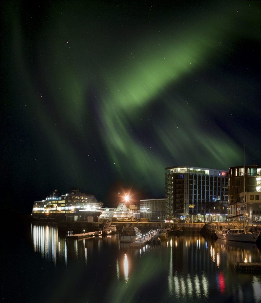 Northern lights over a harbour at night, with a docked cruise ship and illuminated waterfront buildings.