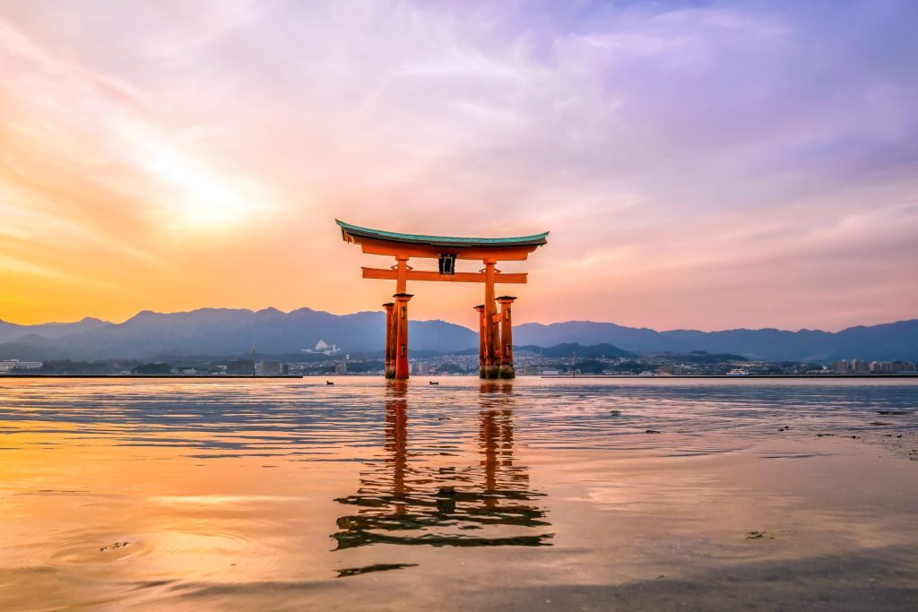 Red torii gate standing in shallow water at sunset, with mountains and a city across the bay.
