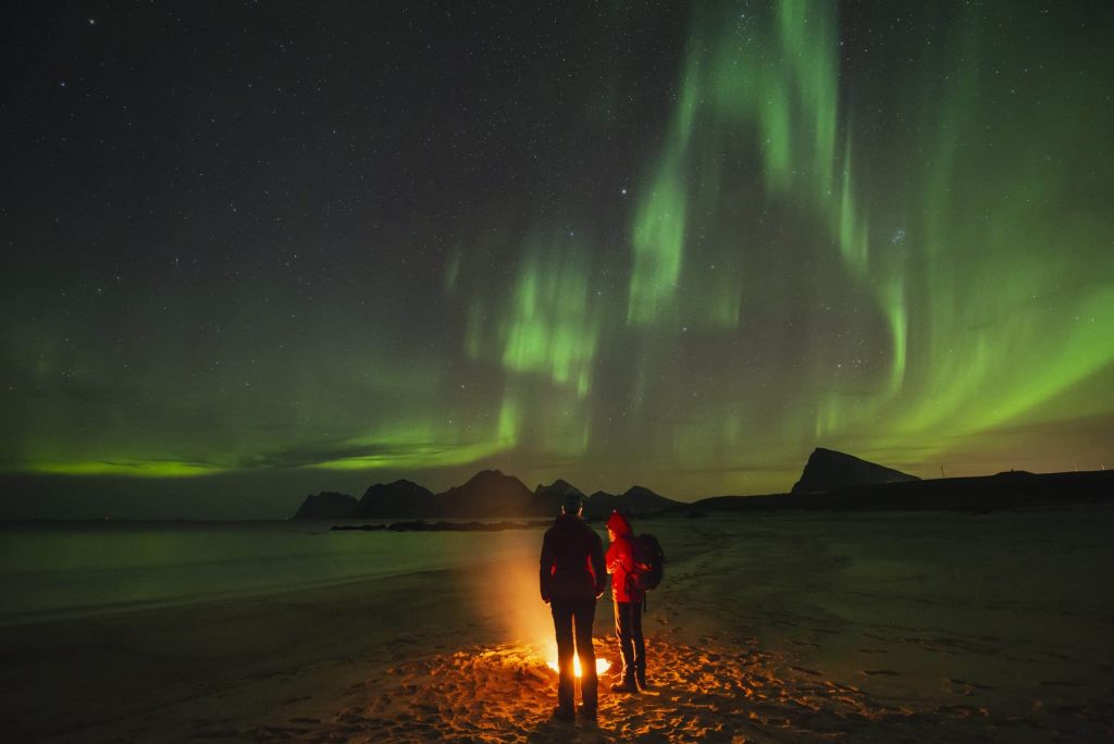 Two people warm themselves by a beach campfire with northern lights – aurora borealis in the sky above, Flakstadøy, Lofoten