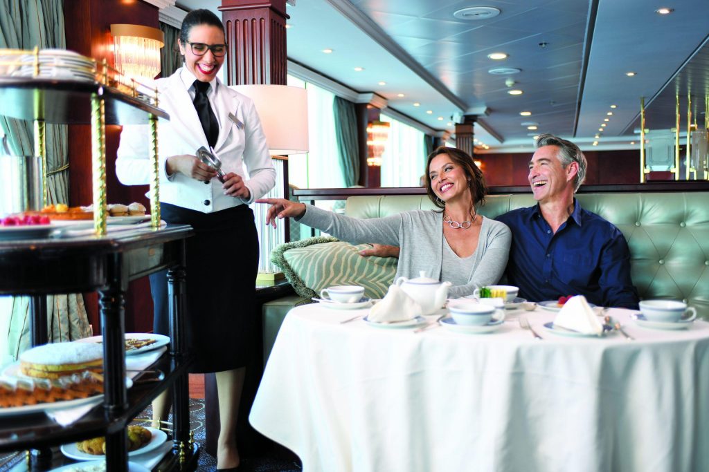 Smiling couple being served afternoon tea and pastries by a waiter in a lounge.
