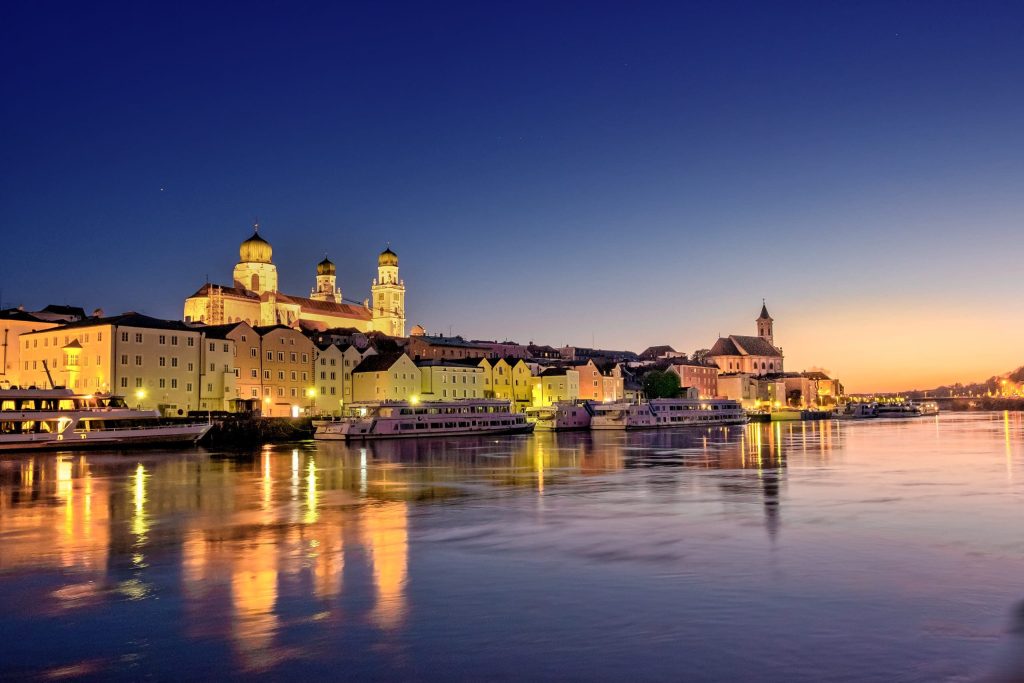 Passau at dusk: floodlit domed cathedral and riverfront buildings reflected in the Danube, with tour boats moored.