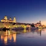 Passau at dusk: floodlit domed cathedral and riverfront buildings reflected in the Danube, with tour boats moored.