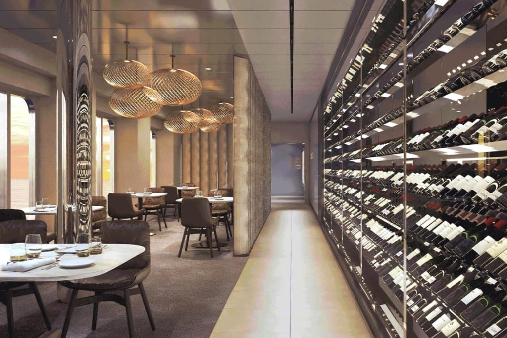 Restaurant with pendant lights and a long wall of wine bottles in a glass-fronted cellar corridor.