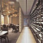 Restaurant with pendant lights and a long wall of wine bottles in a glass-fronted cellar corridor.