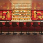 Bar with red and yellow stained-glass cocktail artwork, backlit bottle shelves and dark leather stools.