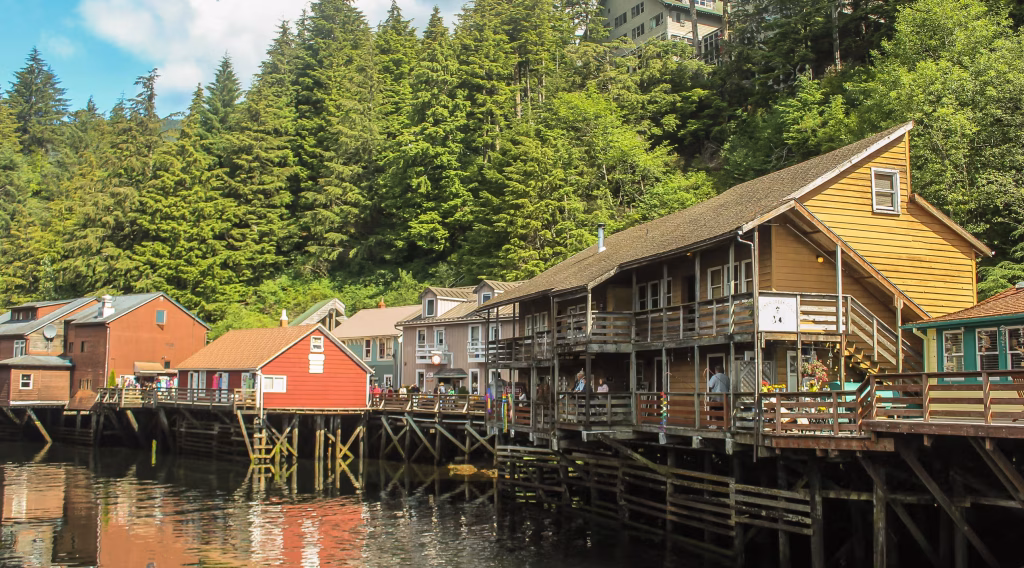 Wooden waterfront buildings on stilts over calm water, backed by dense evergreen forest.