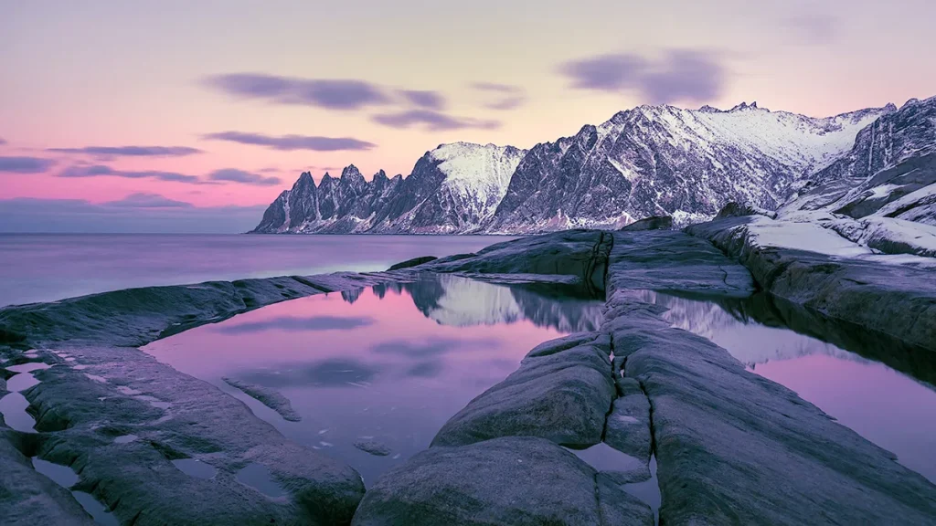 Pink dusk sky over snow-dusted jagged peaks, with rocks and a tidal pool reflecting the mountains.