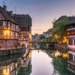Half-timbered houses lining a canal at dusk, with warm lights in the windows and reflections in the water.