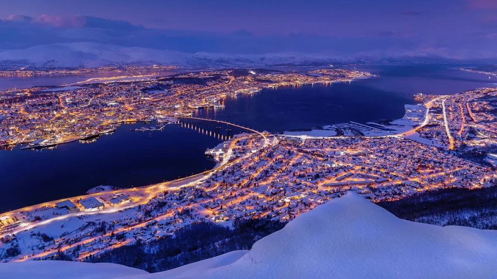 Aerial night view of a glowing city along a fjord and bridge, with snowy mountains in the distance.
