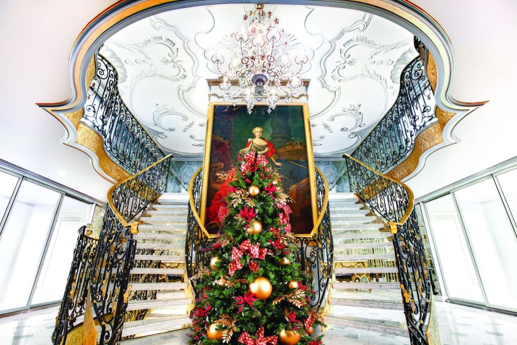 Elegant curved staircase with ornate iron railings, a large framed portrait, and a festive Christmas tree in the center beneath a crystal chandelier.