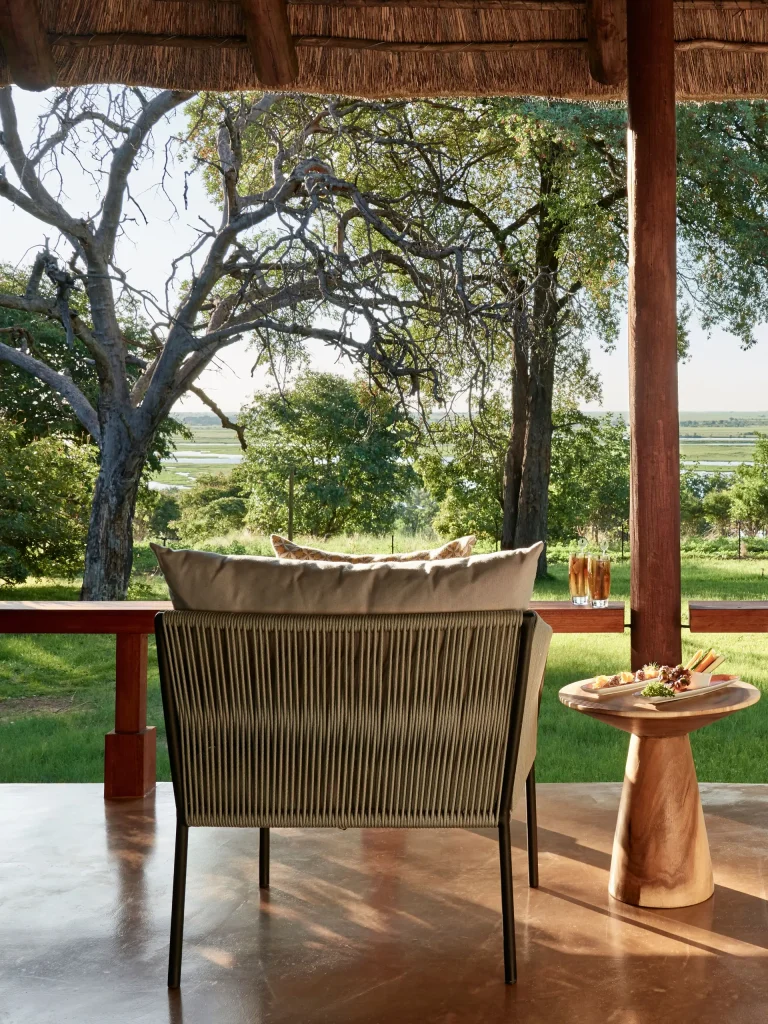 Covered lodge deck with a rope sofa and side table with snacks overlooking trees and grassland.