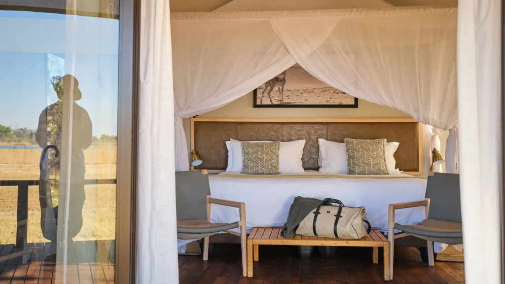 Safari tent bedroom with canopy bed, seating and an open view of grassland and water.