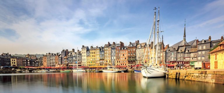 Colorful row of buildings along a harbor with moored sailboats in the foreground.