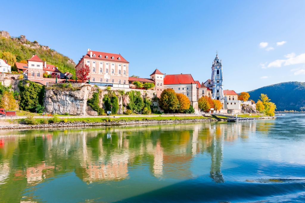 Riverside village with a blue-towered church, pastel buildings and autumn trees along the water.