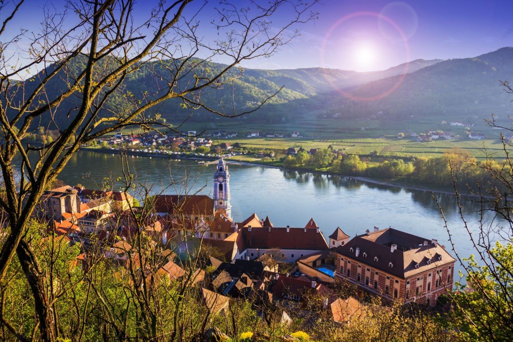 Riverside village with a church tower and red roofs at the edge of a wide river, with vineyards beyond.