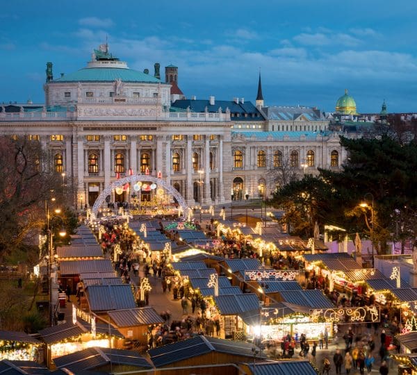 Outdoor Christmas market at dusk with illuminated stalls in front of a grand historic building.
