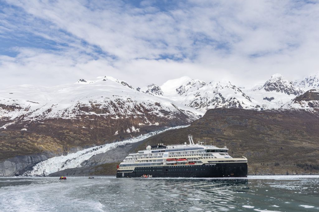 Expedition ship in front of a snow-capped glacier and mountains, with small boats on icy water.
