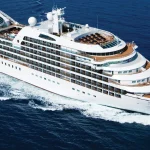 White luxury expedition ship cruising across dark blue sea with a white wake behind it.