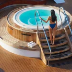 Woman in a blue bikini walking down steps to a round teak-edged hot tub on a ship deck.