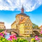 Yellow half-timbered town hall on an arched stone bridge over a river, framed by pink flowers.