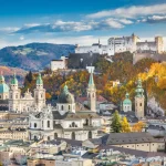 Hilltop castle fortress above a historic city of domed churches, with autumn trees and mountains.