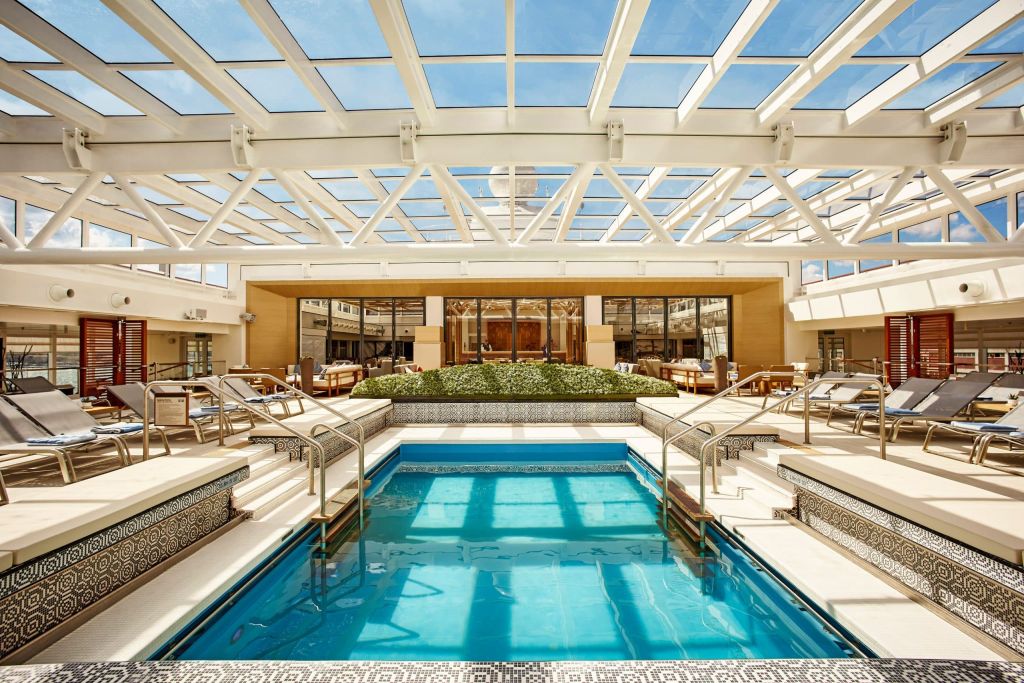 Covered pool with a glass roof, teak loungers and blue-tiled pool water.