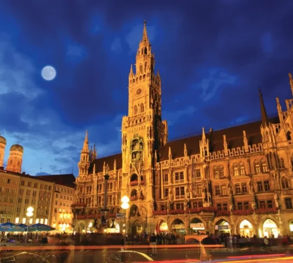 Gothic town hall lit up at night in a busy square, with a full moon in the blue sky.