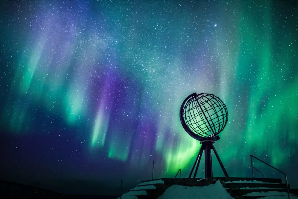 Northern lights over a metal globe monument on a snowy platform at night.