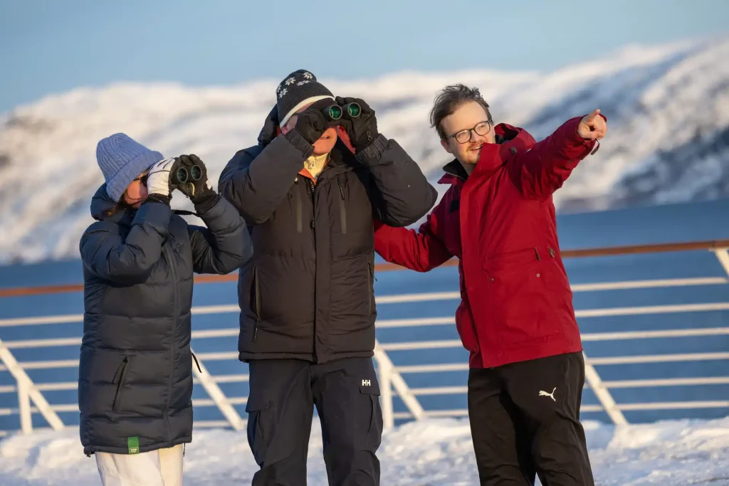 Three people in winter coats and hats look through binoculars on a ship deck with snowy hills behind.