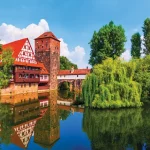 Half-timbered red-roofed houses and a medieval tower beside a canal, with willow trees and reflections in the water.